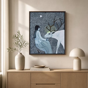 Pode incluir: Uma impressão de arte de parede emoldurada apresentando uma pessoa com cabelo trançado em um vestido claro sentada ao lado de um grande cervo branco com chifres escuros. O fundo é uma superfície escura e texturizada com escrita branca.