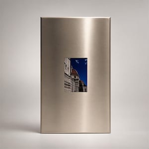 Op de afbeelding: Een rechthoekige wandplaat van geborsteld zilverkleurig metaal met een gecentreerde, verticale rechthoekige uitsparing. Door deze opening is een foto van de koepel van de kathedraal van Florence zichtbaar, met architectonische details tegen een diepblauwe lucht.