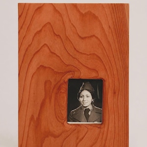 Puede incluir: Un marco de fotos rectangular de madera con un patrón de veta de madera de color marrón rojizo prominente. El marco presenta una superficie lisa y plana y contiene una pequeña fotografía en blanco y negro centrada de una persona con uniforme.