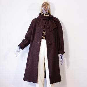May include: A dark brown wool coat featuring a high collar with a side tie detail, long sleeves, and large round buttons down the front. The coat is worn over a brown and black animal print top and plain off-white trousers on a mannequin.