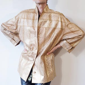 May include: A metallic gold jacket featuring a textured snakeskin pattern throughout. The garment has a high collar, long sleeves with cuffs, and a button-down front closure. The design consists of vertical panels of varying snakeskin textures.