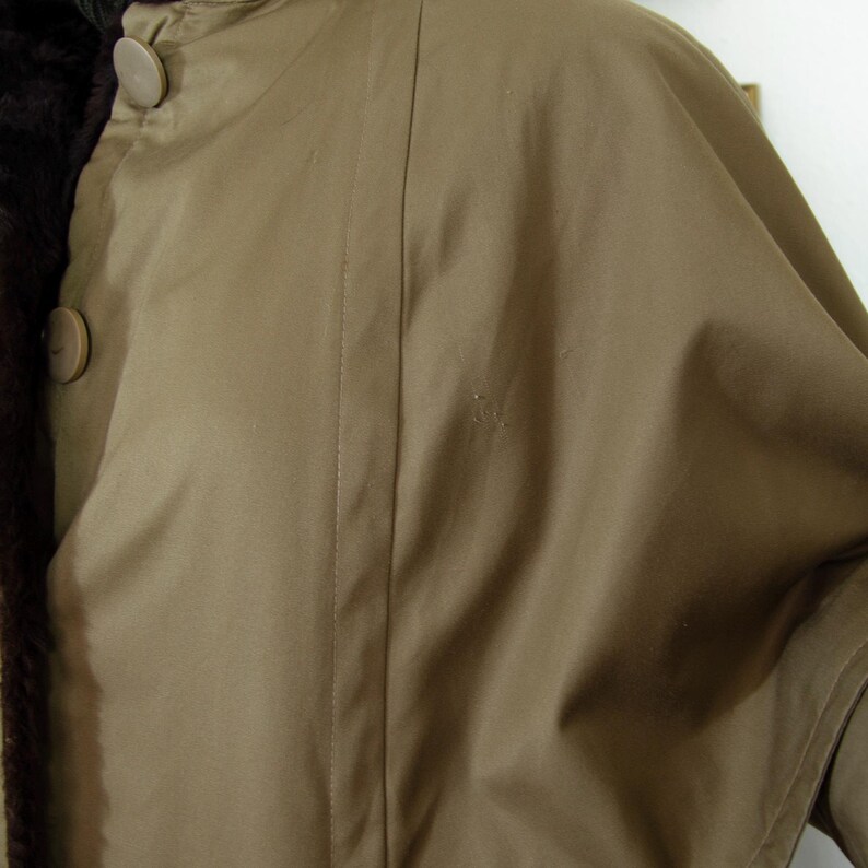 May include: A close-up of a tan-colored coat with a brown fur lining. The coat has a button closure and a cape-like design. The fabric appears smooth and the coat is likely made of a durable material.
