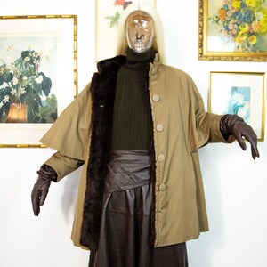 May include: A tan cape coat with a brown faux fur lining and large buttons. The coat is worn over a dark green turtleneck sweater and a brown leather skirt. The mannequin is wearing brown leather gloves.
