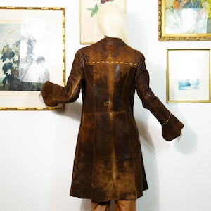 May include: A brown leather coat with a flared skirt and bell sleeves. The coat has a button closure and decorative stitching on the back. The coat is displayed on a mannequin.