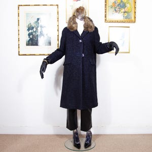 May include: A navy blue wool coat with a faux fur collar, displayed on a mannequin. The coat has a button closure and two front pockets. The mannequin is wearing black gloves, black trousers, and black high heels. Framed artwork is in the background.