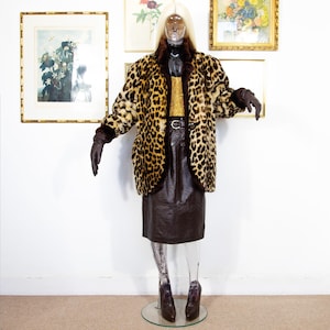 Pode incluir: Um manequim exibe um casaco vintage com estampa de leopardo e acabamentos marrons, combinado com uma saia de couro marrom, um top dourado com lantejoulas e luvas marrons. O conjunto inclui um cinto preto e saltos altos marrons. O fundo apresenta obras de arte emolduradas.