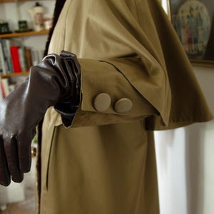 May include: A tan trench coat with a dark brown fur collar and matching brown leather gloves. The coat features two large, round, tan buttons on the sleeve. The coat's fabric has a slight sheen.