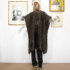 Puede incluir: Una prenda negra y dorada con estampado de capa y cuello redondo, usada sobre pantalones de encaje negros. El atuendo se exhibe en un maniquí transparente con peluca rubia y pendientes dorados. El maniquí está sobre una base de cristal.