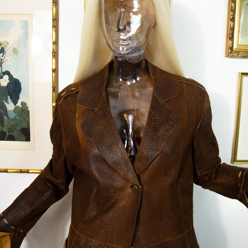 May include: A brown leather blazer with a single-button closure, displayed on a mannequin. The blazer has a textured surface and a classic lapel design. The mannequin has blonde hair and is positioned in front of framed artwork.
