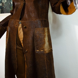 May include: A long, brown leather coat with a textured finish. The coat features two large pockets, button closures, and decorative stitching. The interior lining is a lighter shade of brown.