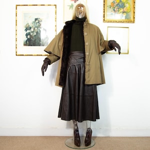 May include: A mannequin displays a vintage olive green cape coat with a brown fur lining and large buttons. The outfit includes a dark green turtleneck sweater, a brown leather skirt, and brown leather gloves. The mannequin is wearing brown leather boots.