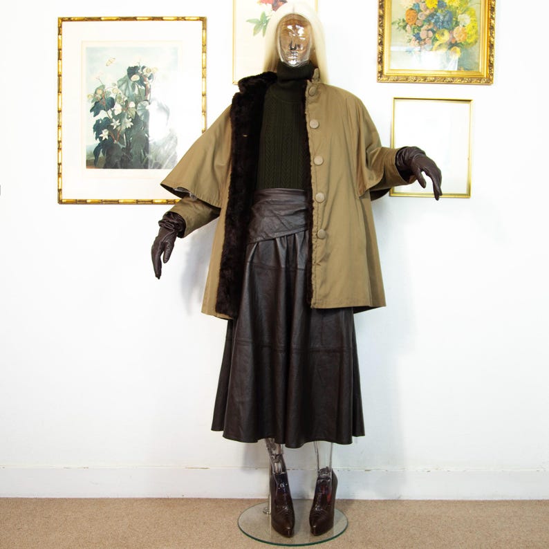 May include: A mannequin displays a vintage olive green cape coat with a brown fur lining and large buttons. The outfit includes a dark green turtleneck sweater, a brown leather skirt, and brown leather gloves. The mannequin is wearing brown leather boots.