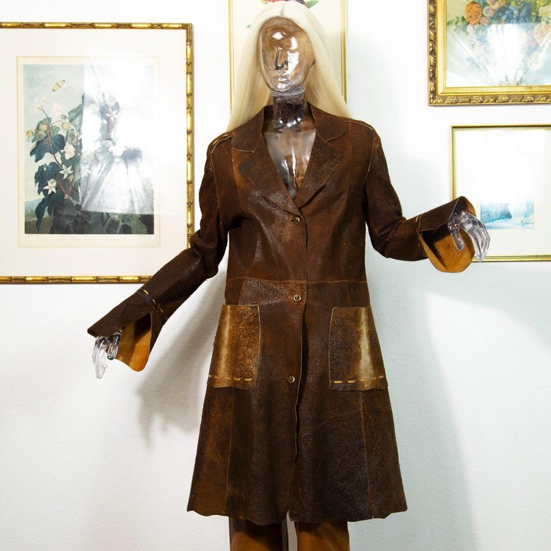 May include: A brown leather coat with a notched collar, two front pockets, and button closures. The coat has a flared skirt and long sleeves with flared cuffs. The coat is displayed on a mannequin with a clear head and hands.