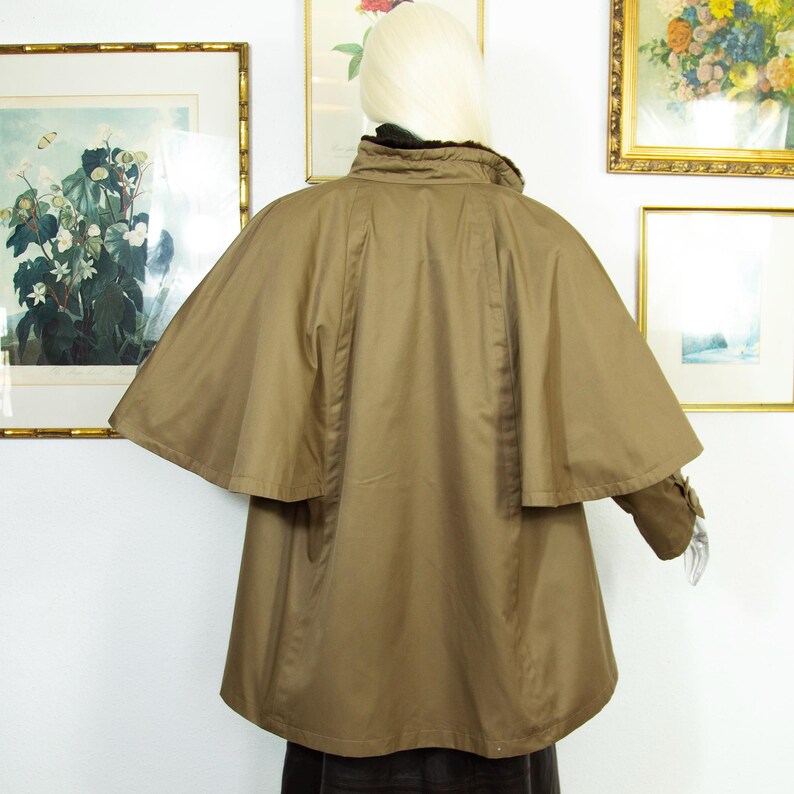 May include: A tan cape coat with a high collar and a layered design. The coat has a buttoned sleeve detail and a dark lining around the collar. The back view shows the cape's flowing shape and tailored seams. The coat is displayed on a mannequin.