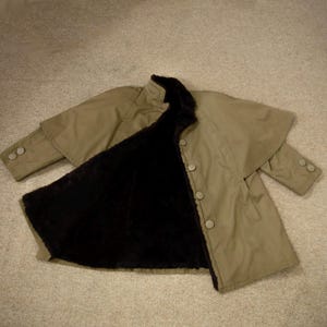 May include: A tan coat with a black faux fur lining. The coat has a cape-like design over the shoulders, long sleeves, and large buttons down the front. The coat is open, revealing the dark interior.