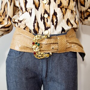 May include: A wide beige snakeskin-textured belt featuring a large, ornate gold-tone buckle. The buckle is shaped like a crescent moon and is embellished with clear rhinestones and three smooth, round cabochon stones in red and green colors.