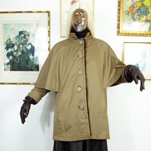 May include: A tan cape coat with three large buttons down the front. The coat has a high collar and a cape-like design over the shoulders. The mannequin is wearing dark brown gloves and a black turtleneck. The background includes framed artwork.