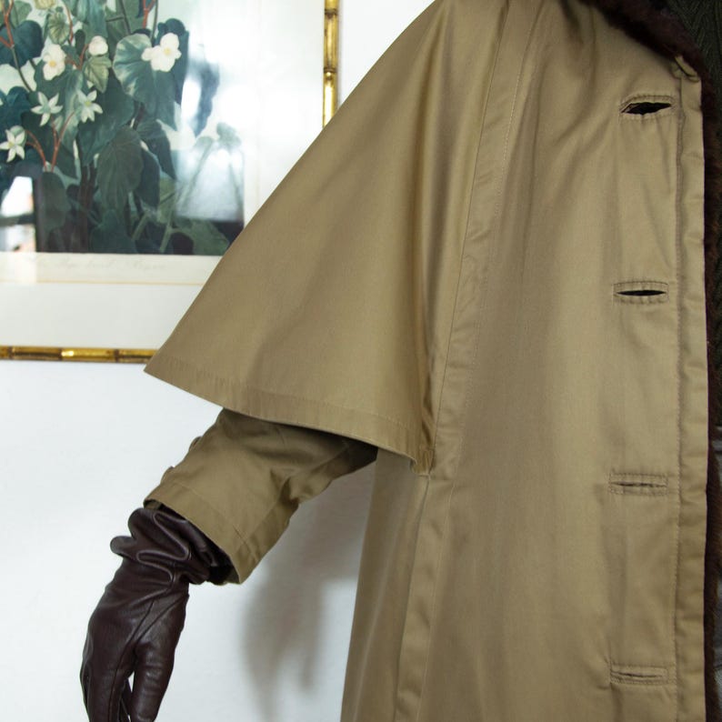 May include: A tan trench coat with a cape-like overlay on the upper sleeves. The coat has three visible buttons and a dark brown fur collar. The sleeve of a brown leather glove is visible.