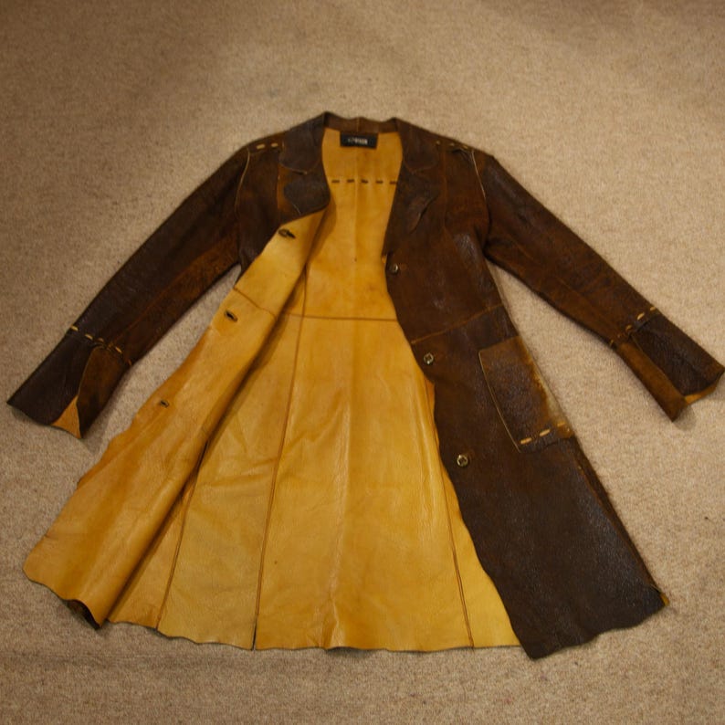 May include: A brown leather coat with a yellow interior, laid flat. The coat has a button closure, pockets, and decorative stitching. The sleeves are long, and the coat appears to be a medium length.