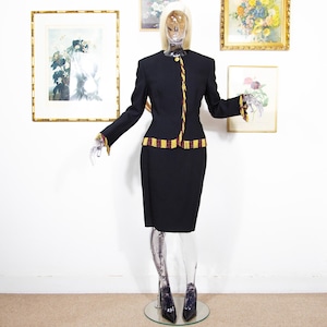 May include: A black skirt suit displayed on a mannequin. The suit features a long-sleeved jacket with gold and burgundy trim and a matching knee-length skirt. The jacket has a single button closure. The mannequin is wearing black pointed-toe heels.