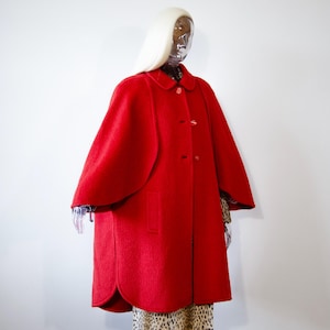 May include: A bright red wool-textured cape coat featuring a rounded collar, three front buttons, and wide arm openings. The garment has a structured silhouette with a curved hemline, extending to the mid-thigh, worn over a leopard print garment.