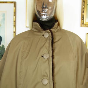 May include: A tan cape-style coat with a high collar and three large, round buttons. The coat has a dark brown fur trim along the edges and a matching lining. The coat is displayed on a mannequin.