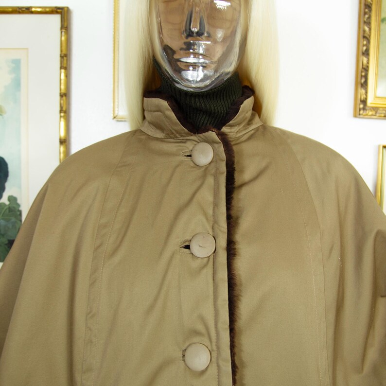 May include: A tan cape-style coat with a high collar and three large, round buttons. The coat has a dark brown fur trim along the edges and a matching lining. The coat is displayed on a mannequin.
