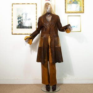 May include: A vintage brown leather coat with a flared skirt and two front pockets. The coat has a notched collar and button closure. The outfit includes matching brown leather pants and brown leather boots. The mannequin has clear plastic hands and a clear plastic head.
