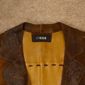 May include: Close-up of a brown leather jacket with a yellow interior. The jacket has a notched collar and a label that reads "BISON". The leather has a distressed appearance, with visible stitching and a textured surface.
