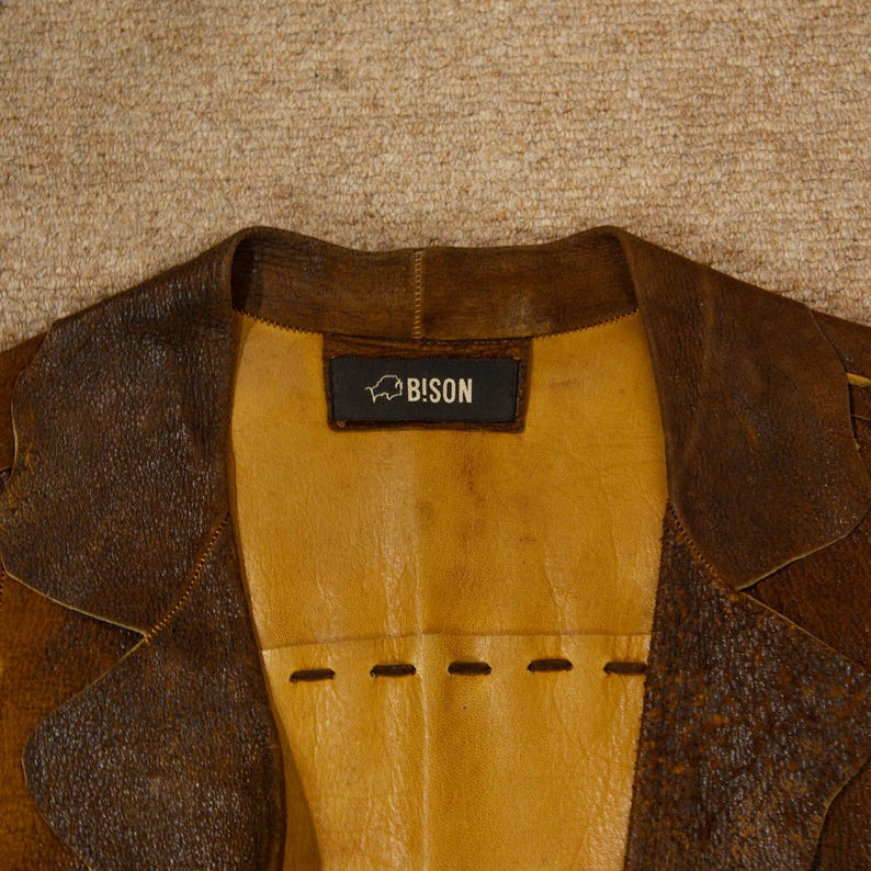 May include: Close-up of a brown leather jacket with a yellow interior. The jacket has a notched collar and a label that reads "BISON". The leather has a distressed appearance, with visible stitching and a textured surface.