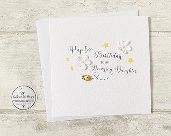May include: A white greeting card with the text "Hapbee Birthday to an Amazing Daughter". The card features white flower and yellow star embellishments, along with a small bee illustration. The card is layered on top of another white card.