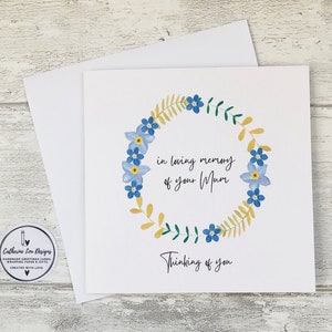 May include: A white card with a floral wreath design, featuring blue flowers, yellow leaves, and green accents. The text reads "in loving memory of your Mum" and "Thinking of you."