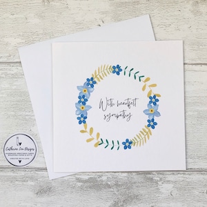 May include: A white sympathy card with a floral wreath design. The wreath features blue flowers and green and yellow leaves. The words "With heartfelt sympathy" are written in black script. An envelope is behind the card.