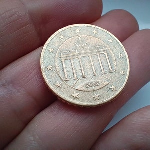 May include: A gold-coloured 1 Euro coin featuring the Brandenburg Gate and the year 2002.