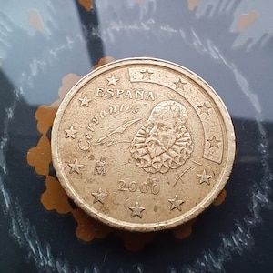 May include: A bronze coin with the inscription "ESPAÑA Cervantes" and the year 2000. It features a portrait of Miguel de Cervantes and is encircled by stars. The coin rests on a dark surface.
