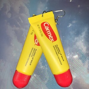 Könnte beinhalten: Ein Paar ausgefallene Ohrringe aus Miniatur-Carmex-Lippenbalsam-Tuben. Die gelben Tuben haben rote Kappen und das Carmex-Logo in Rot. Der Text "CLASSIC LIP BALM" und "NET WT .35 OZ. (10g)" sind sichtbar.