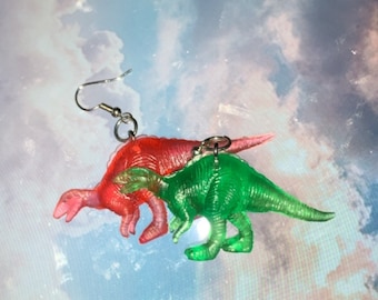 Könnte beinhalten: Ein Paar Ohrringe mit einem grünen und einem rosa Dinosaurier-Anhänger. Die Dinosaurier sind durchsichtig und haben eine glänzende Oberfläche.