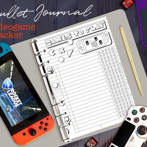 Könnte beinhalten: Ein Bullet Journal mit dem Text "Videogame Tracker" und "Games to Play". Eine Nintendo Switch-Konsole mit dem Spiel Rocket League befindet sich neben dem Journal. Ein goldener Stift und ein weißer Gamecontroller sind ebenfalls sichtbar.
