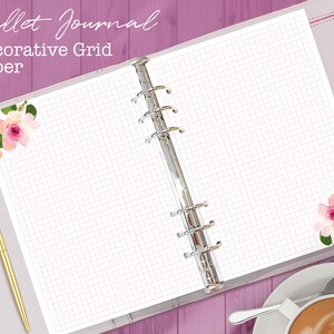 Könnte beinhalten: Ein weißes Notizbuch mit dekorativem Rasterpapier und einem silbernen Ringmechanismus. Das Notizbuch hat an den Ecken der Seiten rosa florale Designs. Der Text "Bullet Journal Decorative Grid Paper" befindet sich oben auf der Seite.