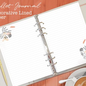 May include: A white, lined, decorative paper notebook with a silver binder. The notebook has a floral design with pink and orange accents on the front and back covers. The text "Bullet Journal Decorative Lined Paper" is printed on the front cover.