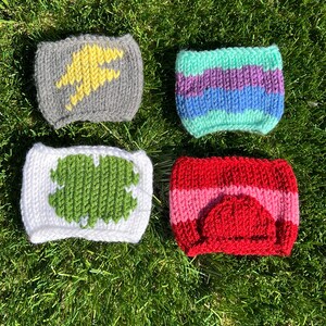 May include: Four knitted cup cozies in different colors and patterns. The first is gray with a yellow lightning bolt design. The second is striped in green, purple, and blue. The third is white with a green clover design. The fourth is red with a pink stripe and a red heart design.