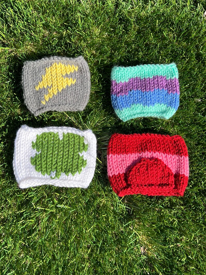 May include: Four knitted cup cozies in different colors and patterns. The first is gray with a yellow lightning bolt design. The second is striped in green, purple, and blue. The third is white with a green clover design. The fourth is red with a pink stripe and a red heart design.