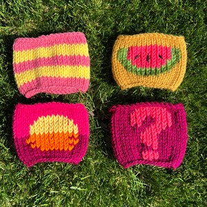 May include: Four knitted cup cozies with different designs. One is pink and yellow stripes, one is yellow with a watermelon design, one is pink with a yellow and orange sun design, and one is pink with a pink question mark.