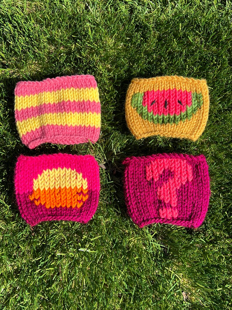May include: Four knitted cup cozies with different designs. One is pink and yellow stripes, one is yellow with a watermelon design, one is pink with a yellow and orange sun design, and one is pink with a pink question mark.