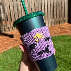 May include: Purple knitted coffee cup cozy with a yellow flower and black eyes. The cozy is on a dark green reusable coffee cup with a green lid and a straw.