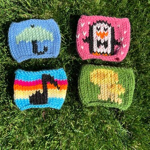 May include: Four knitted cup cozies with different designs. The cozies are blue with a green umbrella, pink with a white penguin, green with a yellow dinosaur, and rainbow striped with a black musical note.