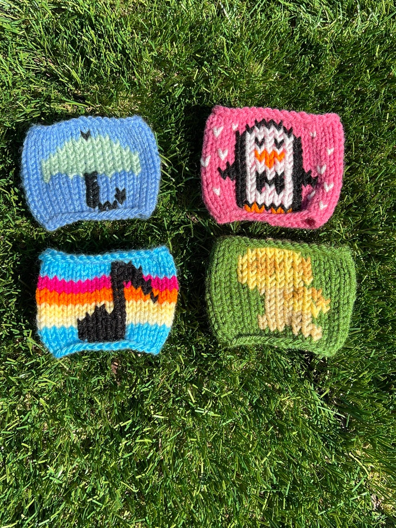 May include: Four knitted cup cozies with different designs. The cozies are blue with a green umbrella, pink with a white penguin, green with a yellow dinosaur, and rainbow striped with a black musical note.