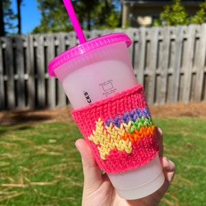 May include: A pink reusable cup with a pink lid and straw. The cup has a knitted pink cup cozy with a rainbow star design. The text "Decaf / Deca" and "Shots / Dosers" is printed on the cup.