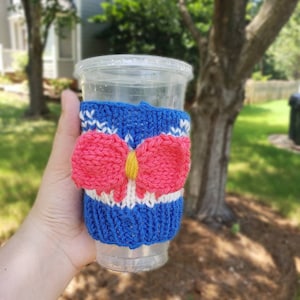 May include: A blue and white knitted cup cozy with a pink bow and yellow center. The cozy is on a clear plastic cup.