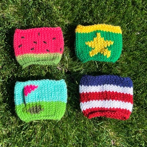 May include: Four knitted cup cozies with different designs. The first is watermelon, the second is a green and yellow star, the third is a golf course, and the fourth is red, white, and blue stripes.
